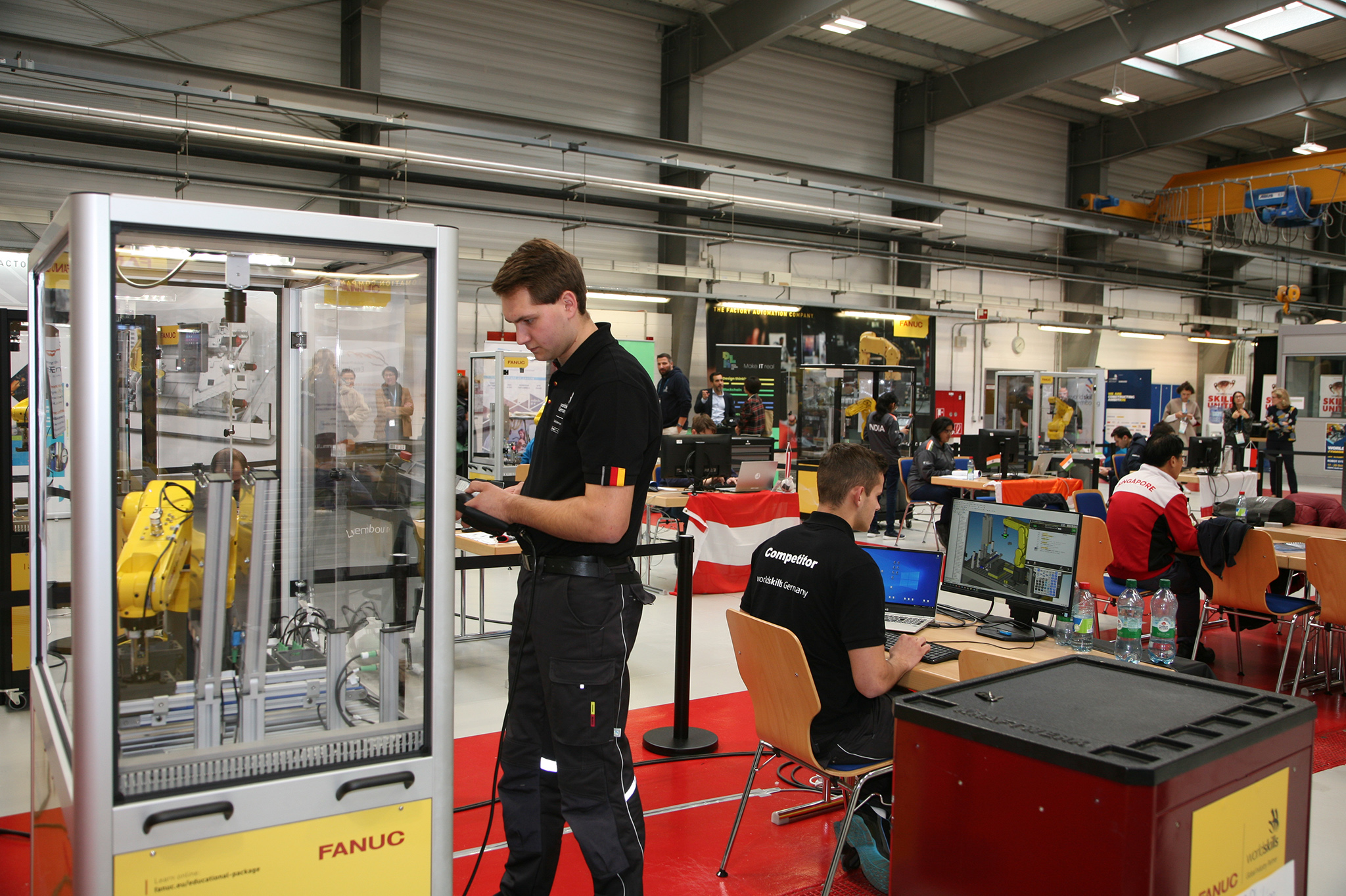 Marvin Schuster standing in front of the robot cell. Philipp Raab sitting at desk and writing codes.